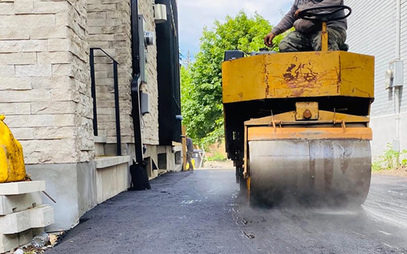 Seal Canada A yellow road roller compacts fresh asphalt for an asphalt repair between two buildings on a narrow alleyway, with a worker operating the machine. Asphalt & Coatings