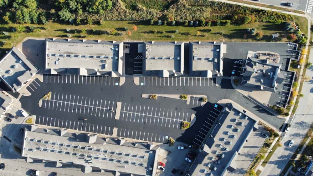 Seal Canada Aerial view of a commercial parking lot with several adjacent buildings and many empty spaces, showcasing recent asphalt repair, surrounded by greenery and roads. Asphalt & Coatings