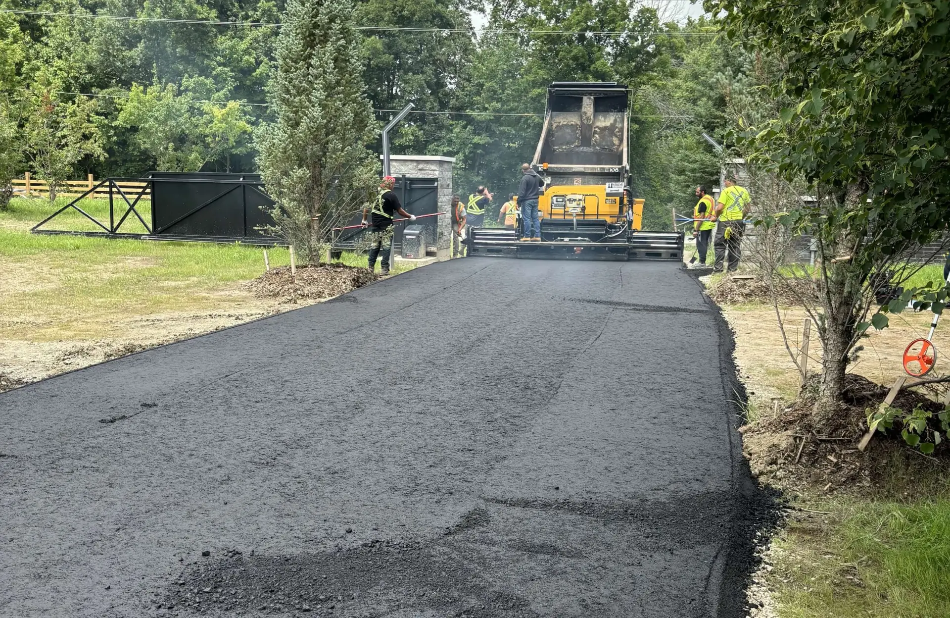 Seal Canada Workers use heavy machinery to lay fresh asphalt on a driveway surrounded by trees and a black gate. Asphalt & Coatings