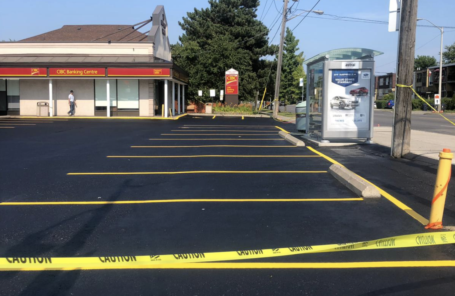 Seal Canada A freshly paved, empty parking lot with yellow caution tape blocking entry, located beside a bank and a bus shelter on a sunny day. Asphalt & Coatings