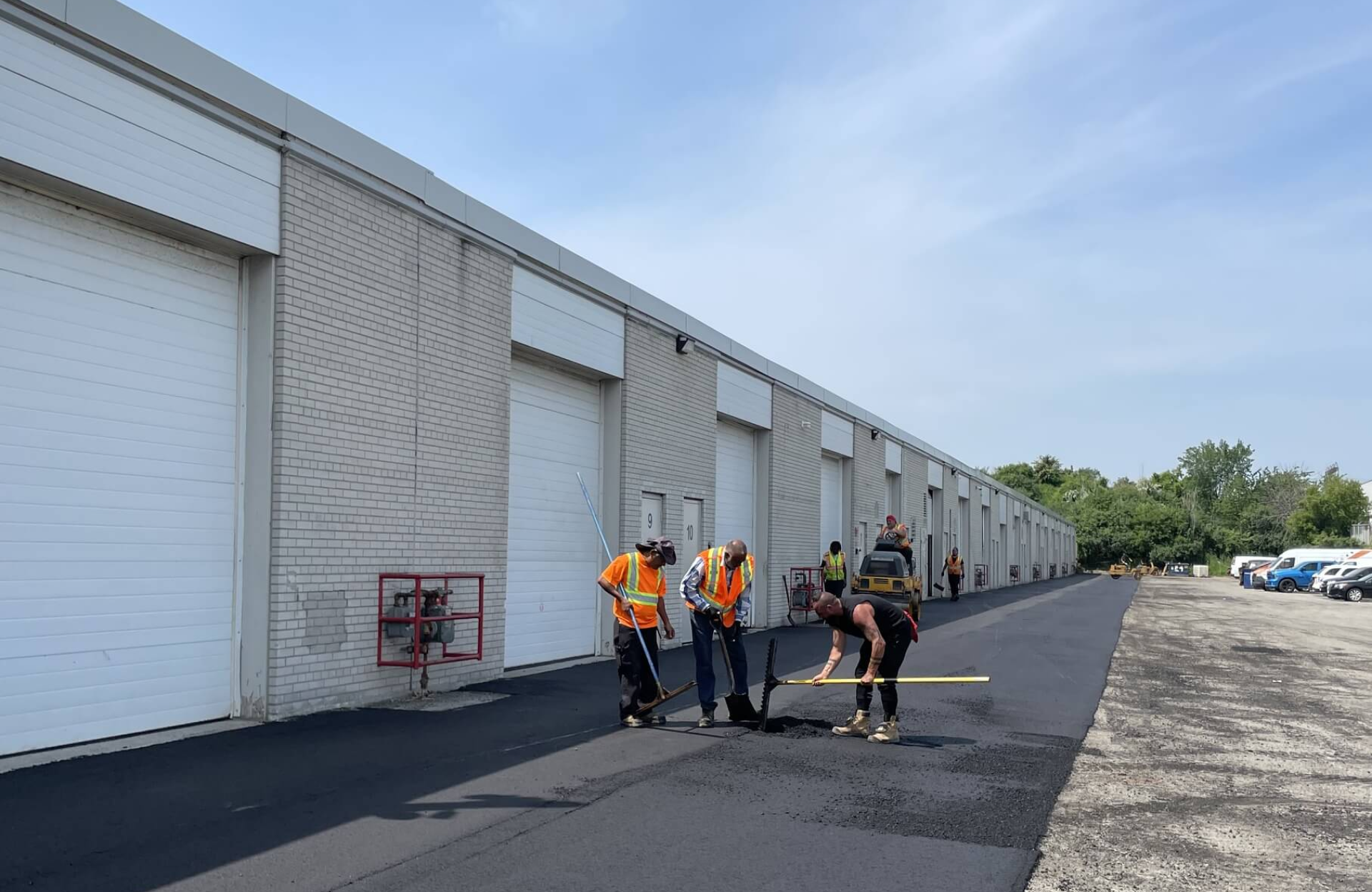 Seal Canada Three workers in safety vests are resurfacing asphalt outside a row of white industrial garages on a clear day, with parked vehicles in the background. Asphalt & Coatings
