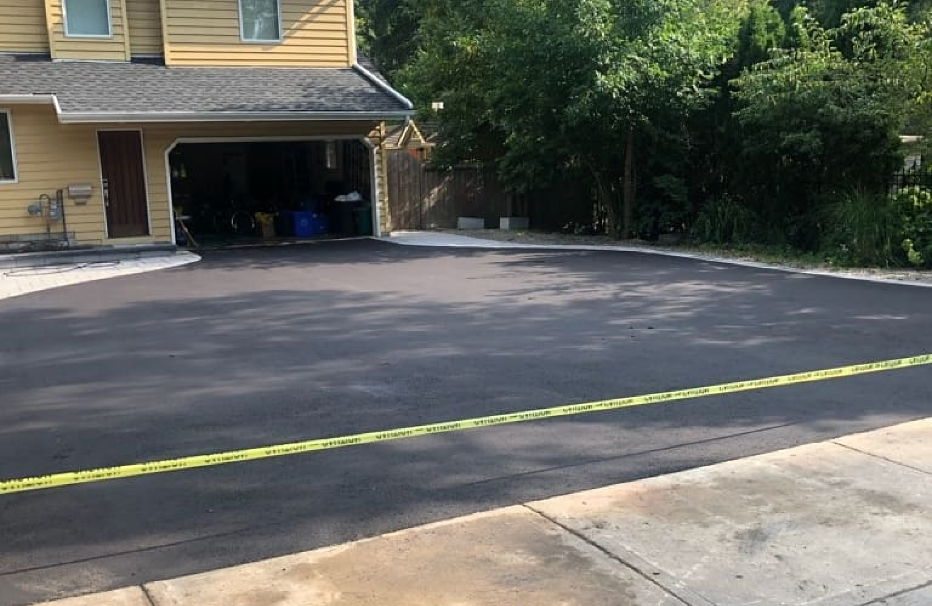 Seal Canada A freshly paved asphalt driveway in front of a garage, blocked off by yellow caution tape, with trees and shrubs in the background. Asphalt & Coatings