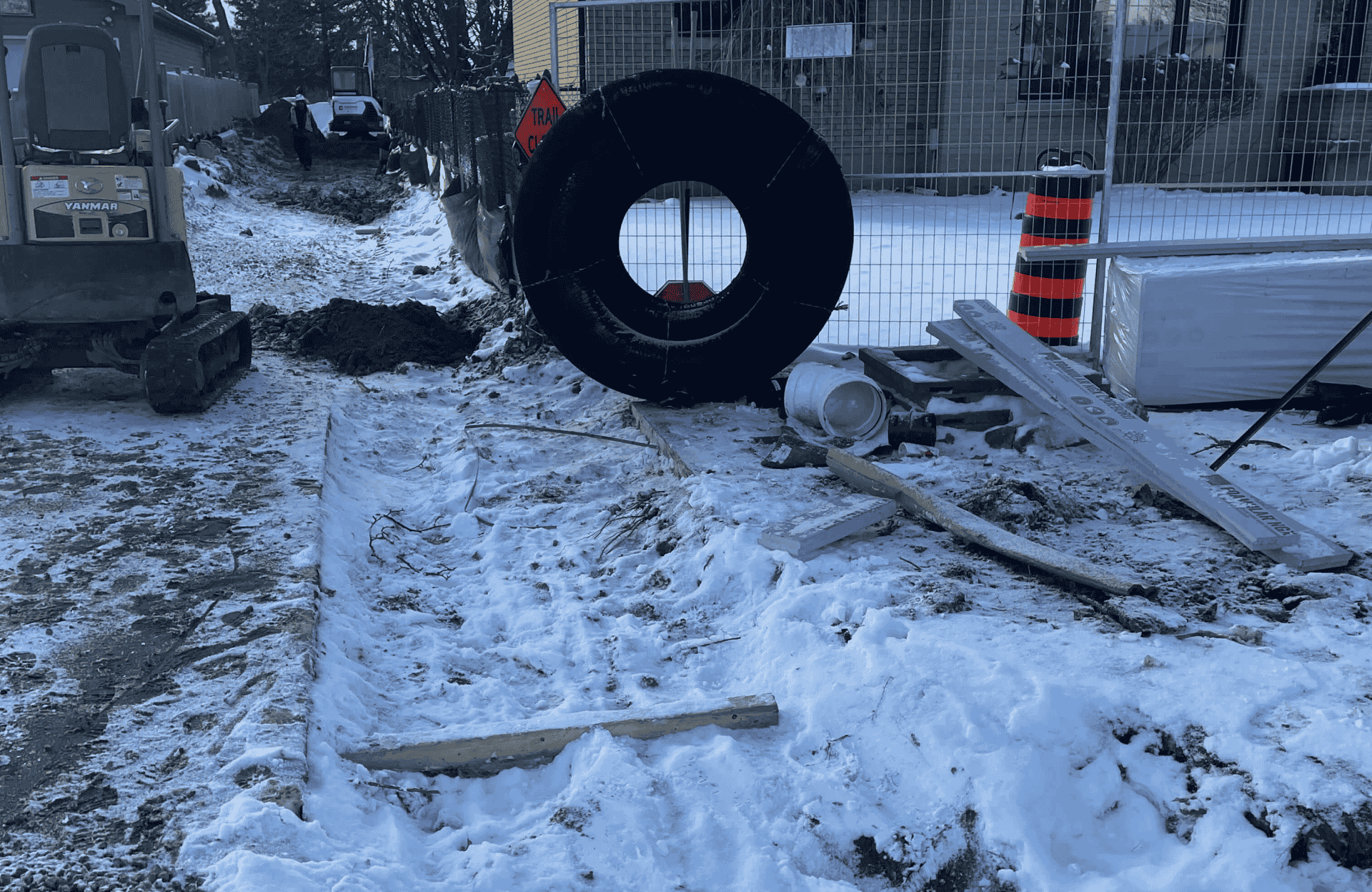 Seal Canada A snow-covered construction site with scattered materials, a large black coil, an excavator on the left, and safety barriers in the background. Asphalt & Coatings