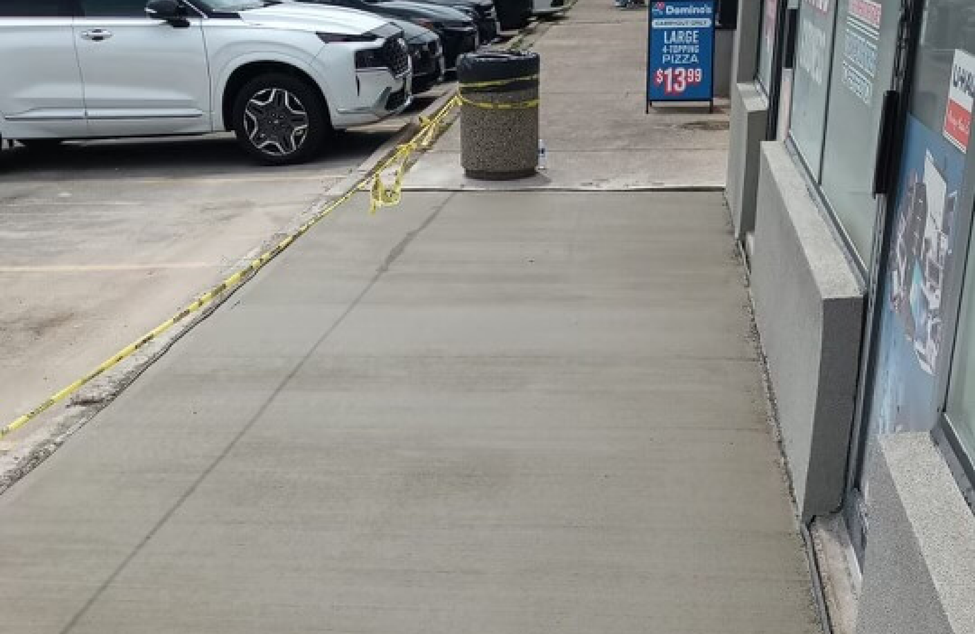 Seal Canada Freshly poured concrete sidewalk in front of a building, bordered by yellow caution tape and a trash can; parked cars and a pizza advertisement are visible in the background. Asphalt & Coatings