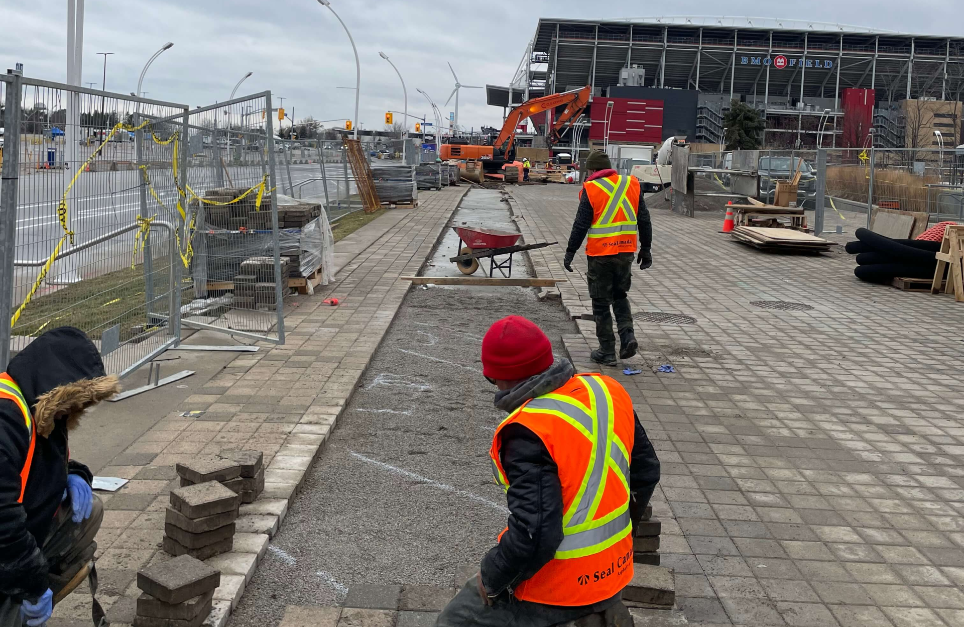 Seal Canada Workers wearing safety vests and hats are repairing a sidewalk with construction equipment and materials near a fenced-off area and a stadium in the background. Asphalt & Coatings