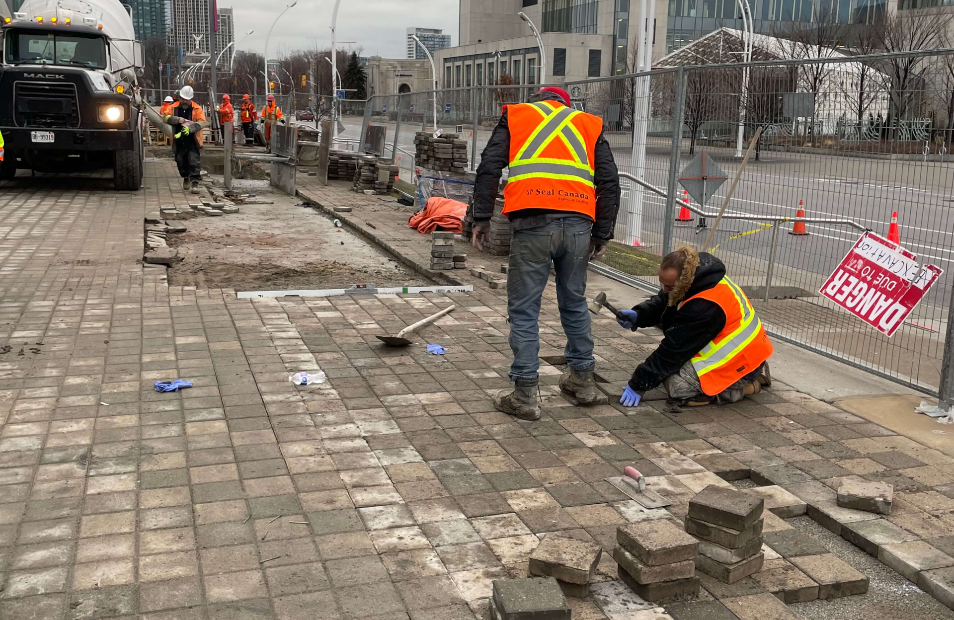 Seal Canada Construction workers in safety vests repair a stone sidewalk, with tools and bricks scattered nearby and fencing blocking off the area. Asphalt & Coatings