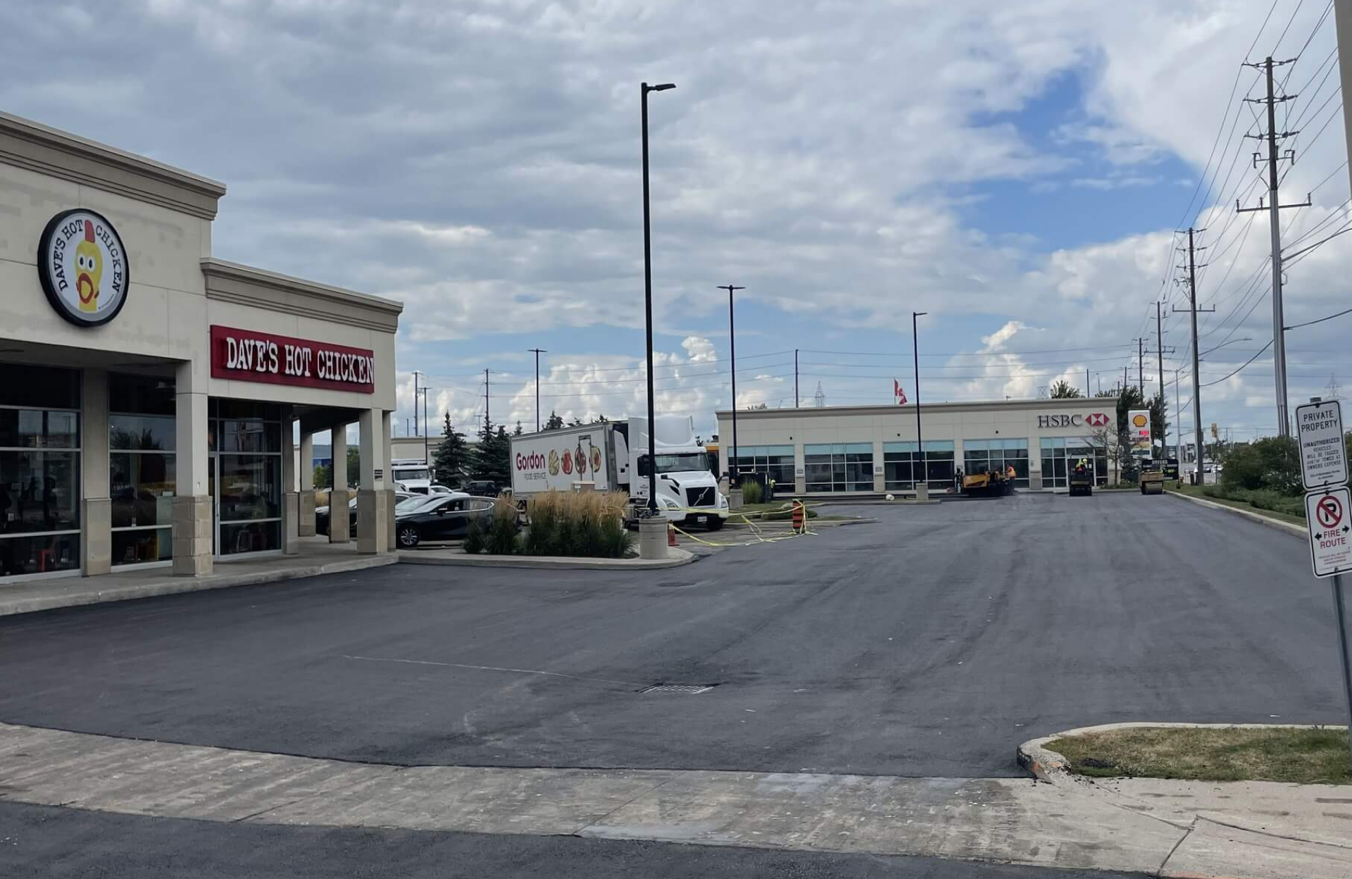 Seal Canada A small shopping plaza with a Dave’s Hot Chicken restaurant on the left and an HSBC bank on the right, separated by a newly paved parking lot under a mostly cloudy sky. Asphalt & Coatings