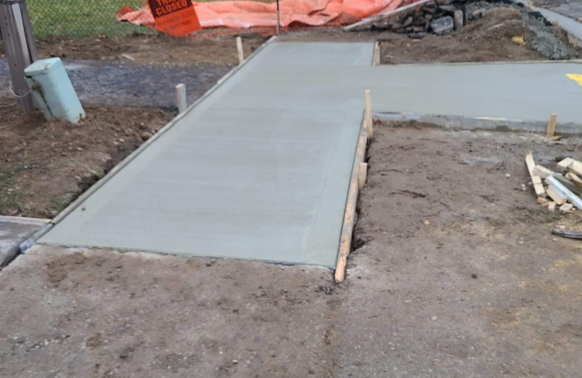 Seal Canada Freshly poured concrete sidewalk with wooden forms still in place, surrounded by dirt and construction materials. Orange construction barrier visible in the background. Asphalt & Coatings