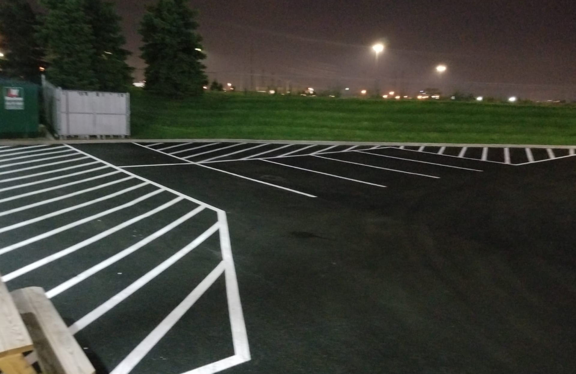 Seal Canada Empty parking lot at night with freshly painted white parking lines, bordered by grass, trees, a dumpster, and illuminated by streetlights in the background. Asphalt & Coatings