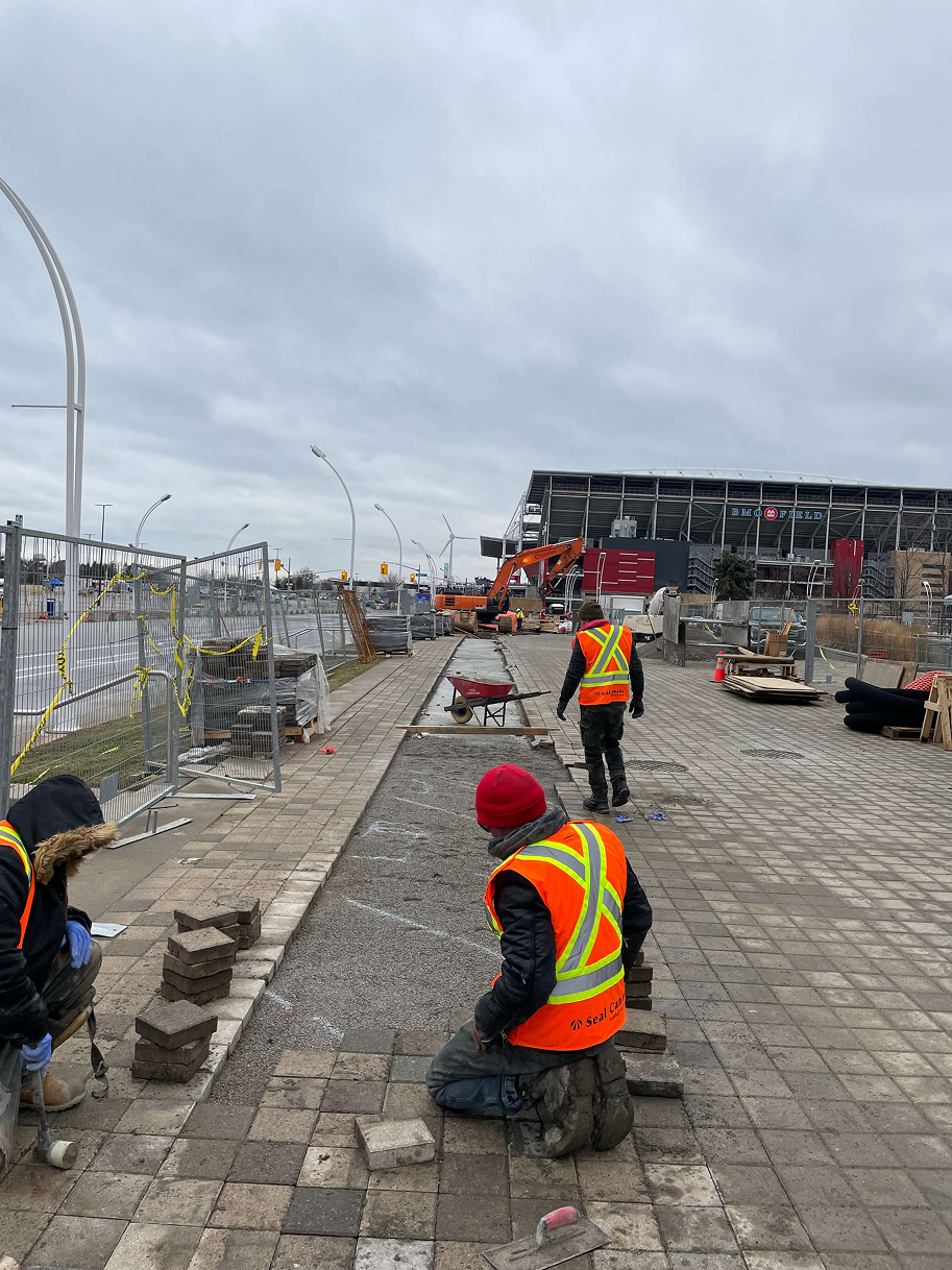Seal Canada Workers wearing safety vests and helmets are laying paving stones on a sidewalk construction site under a cloudy sky, with equipment and materials visible around them. Asphalt & Coatings