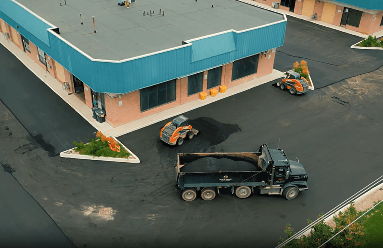 Seal Canada Aerial view of a commercial building with fresh asphalt paving; a dump truck and two compactors are working in the parking lot. Asphalt & Coatings