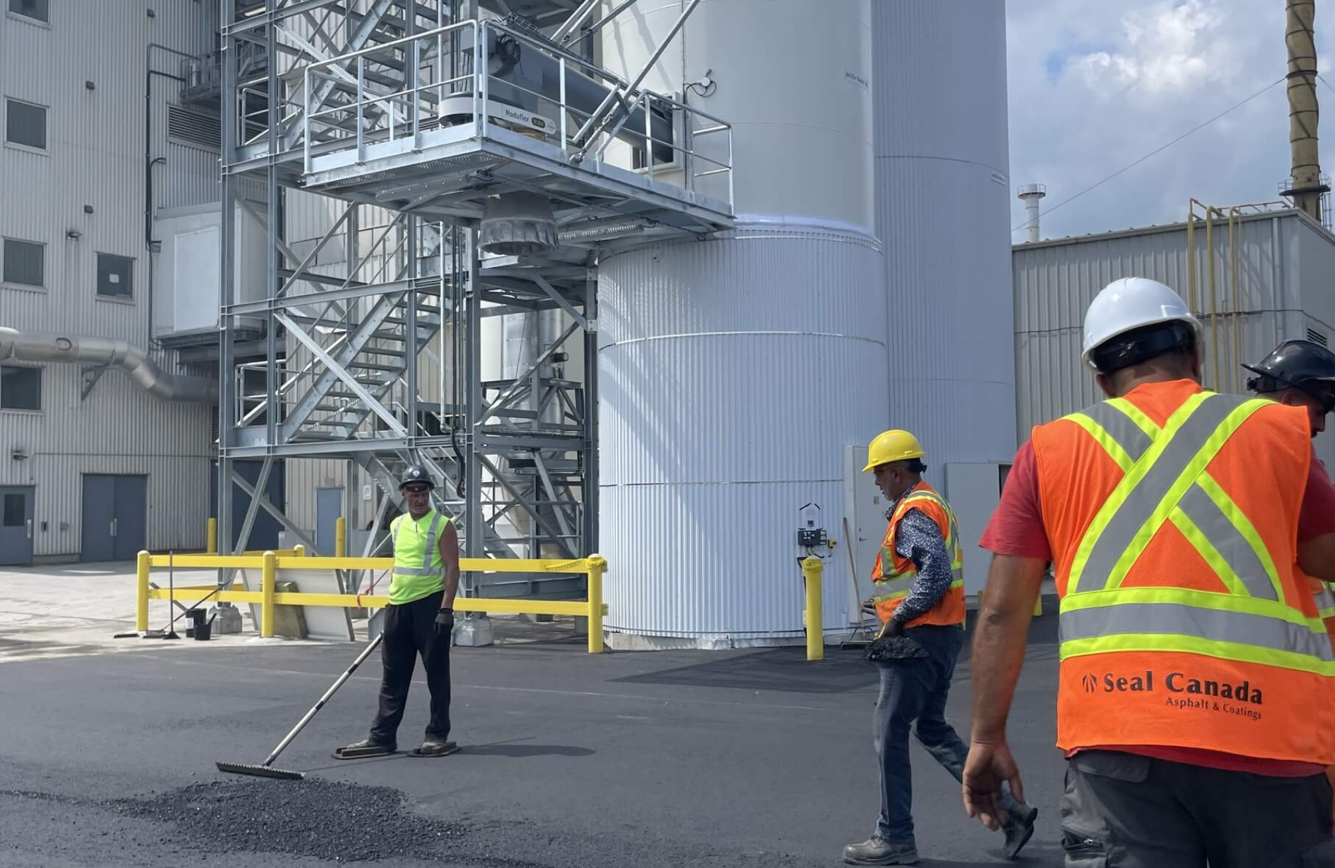 Seal Canada Three construction workers in safety vests and helmets are spreading asphalt on the ground near large industrial storage tanks. Asphalt & Coatings