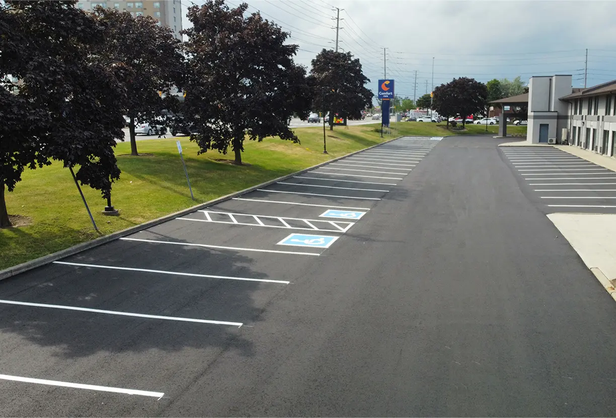 Seal Canada Freshly paved parking lot with clearly marked spaces, including two accessible parking spots near a building, bordered by trees and grass. Asphalt & Coatings