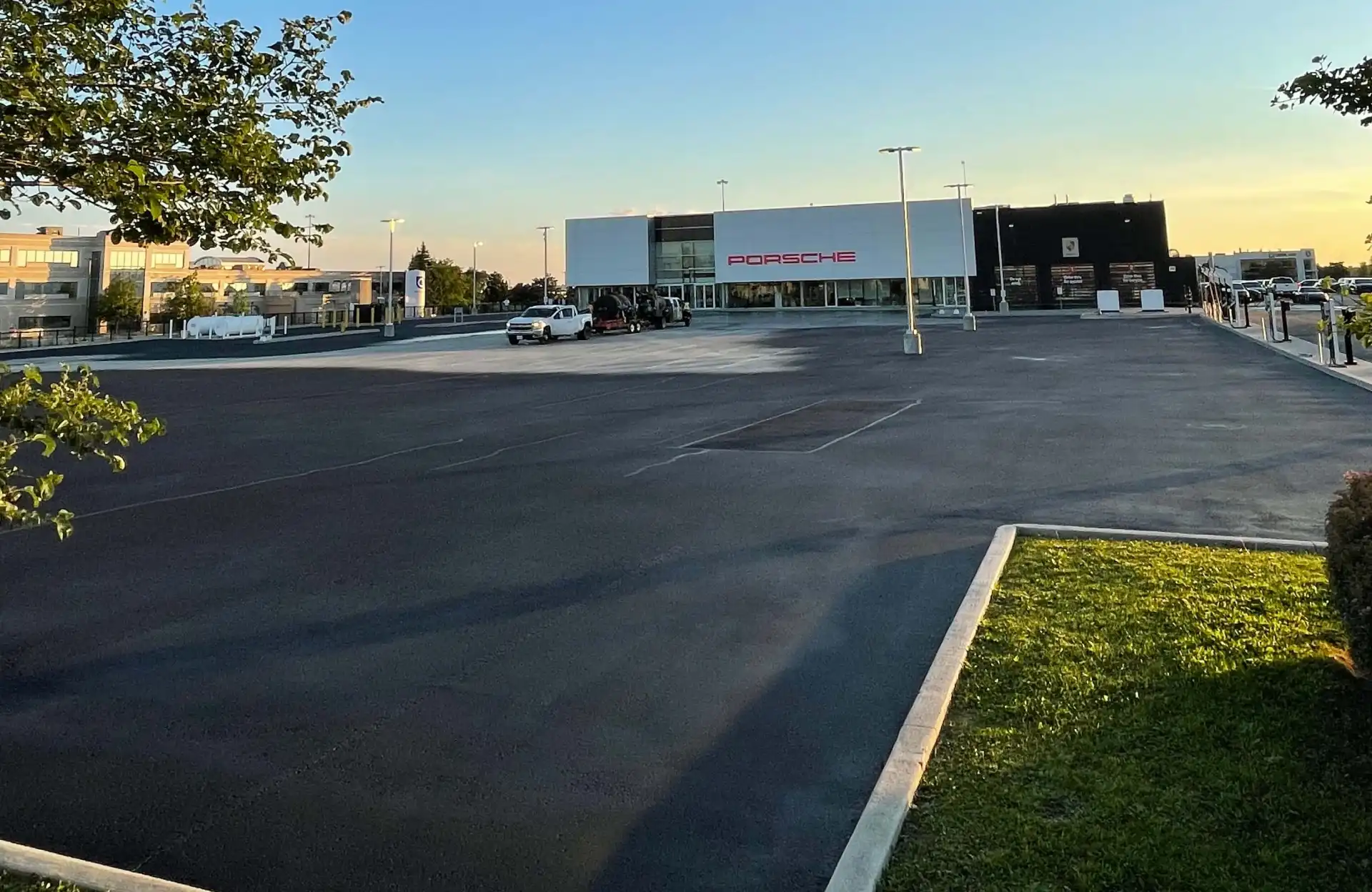 Seal Canada A mostly empty parking lot in front of a Porsche dealership during daylight, with a few cars parked near the building and clear skies overhead. Asphalt & Coatings