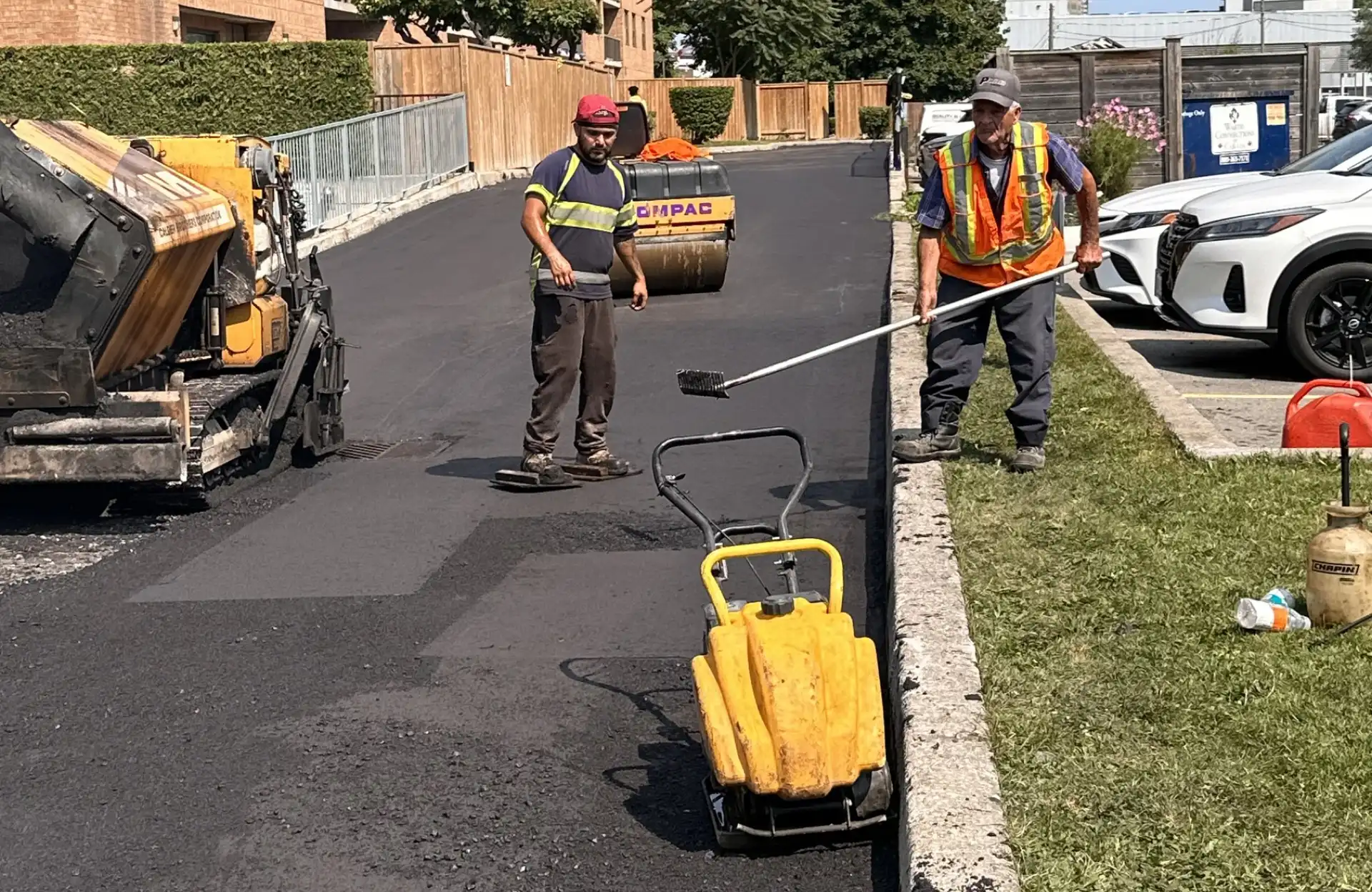 Seal Canada Two workers in safety gear pave an asphalt driveway with construction equipment on a sunny day, near parked cars and a grassy area. Asphalt & Coatings
