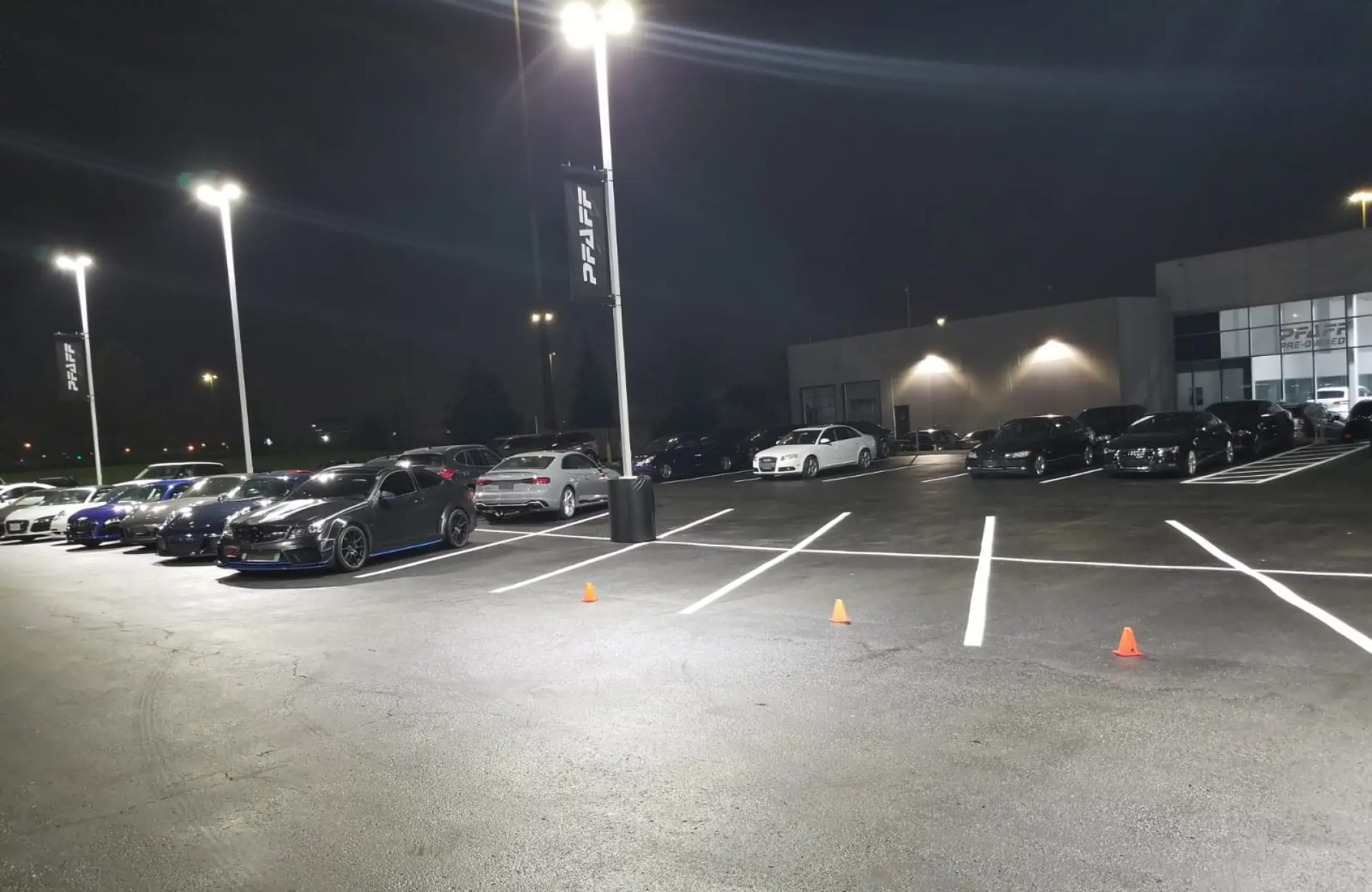 Seal Canada A car dealership lot at night with several parked cars under bright lights; two orange traffic cones are placed in marked parking spaces. Asphalt & Coatings