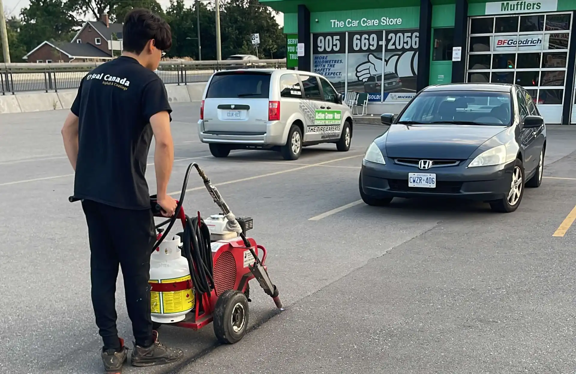 Seal Canada A person operates a wheeled cleaning machine in a parking lot near parked cars outside an automotive service store. Asphalt & Coatings