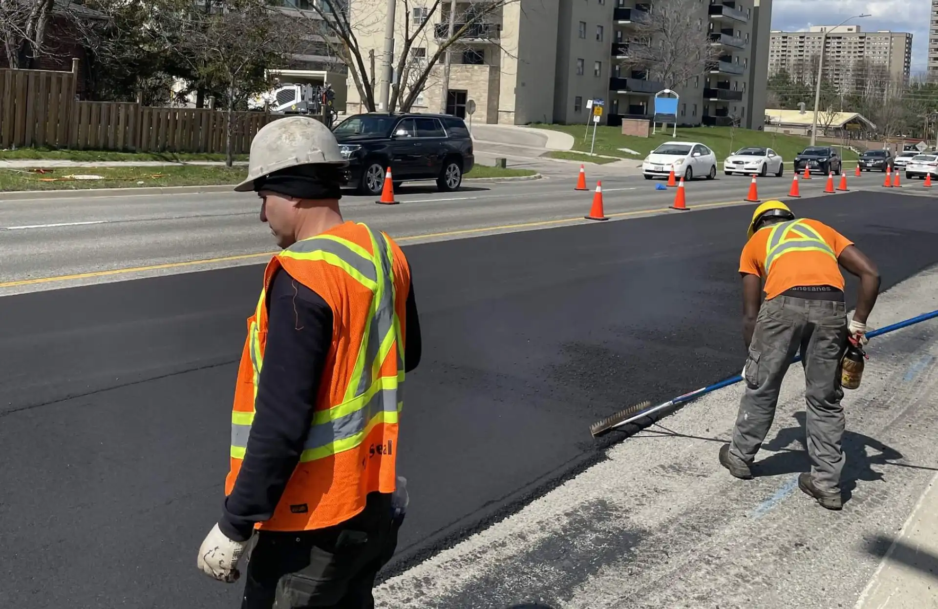 Seal Canada Two construction workers in safety vests resurface a road with fresh asphalt, while traffic cones separate them from passing cars on a sunny day. Asphalt & Coatings