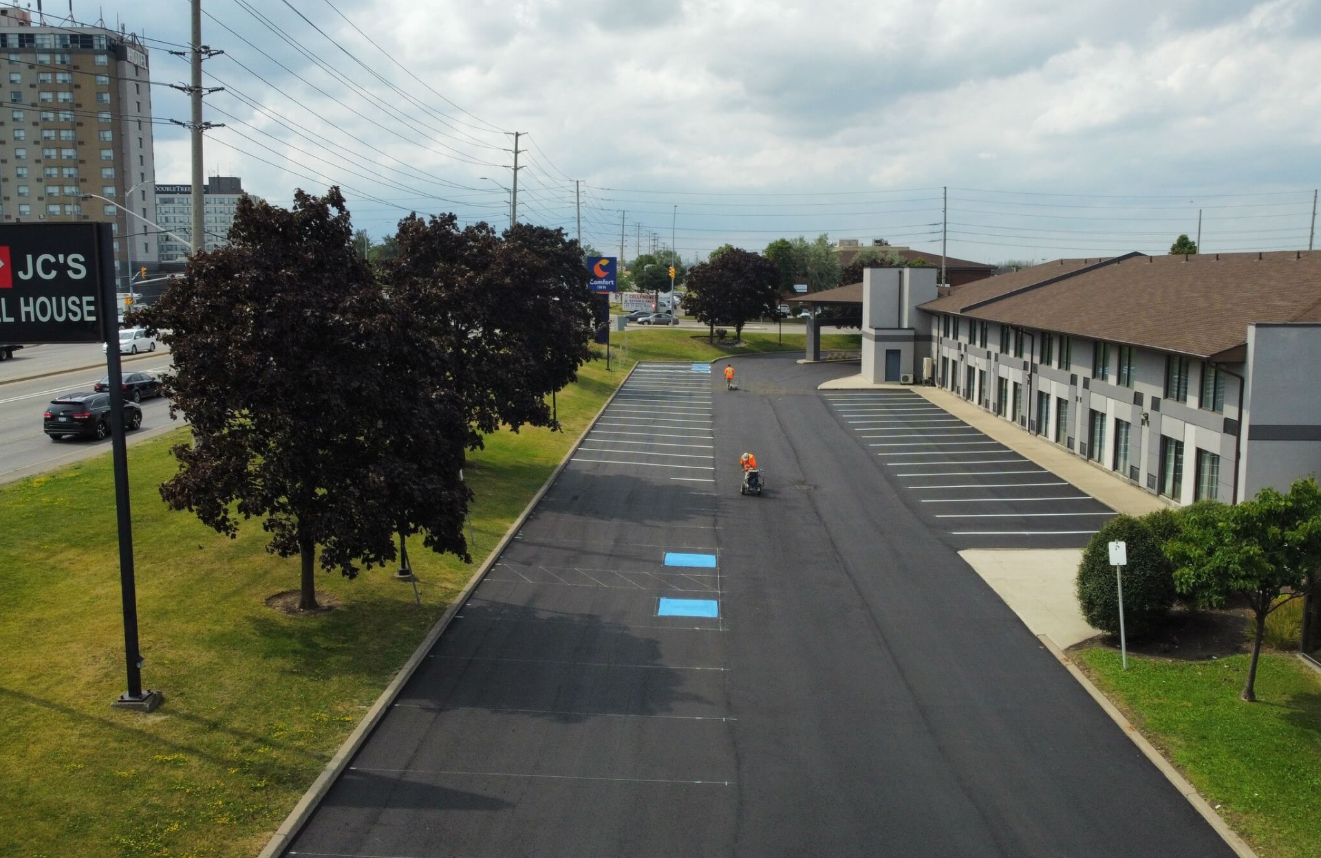 Seal Canada A freshly paved parking lot beside a commercial building, with workers in orange vests painting blue handicap spots under a cloudy sky—demonstrating the same attention to detail found in professional sealcoating for townhouse complexes. Asphalt & Coatings