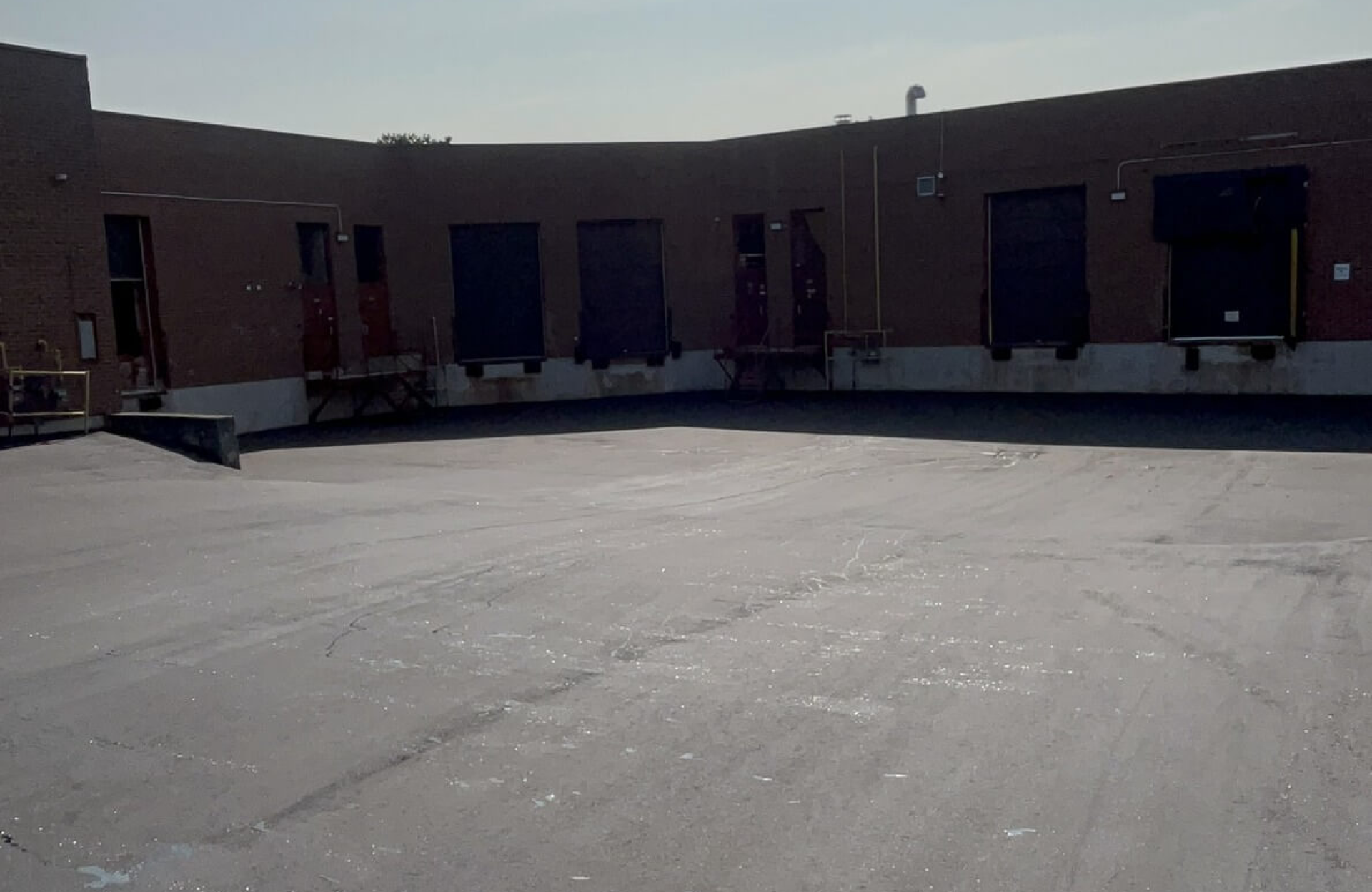 Seal Canada Concrete loading dock area with multiple closed overhead doors on a brick industrial building under a clear sky, showing oil stains on the commercial parking lot surface. Asphalt & Coatings