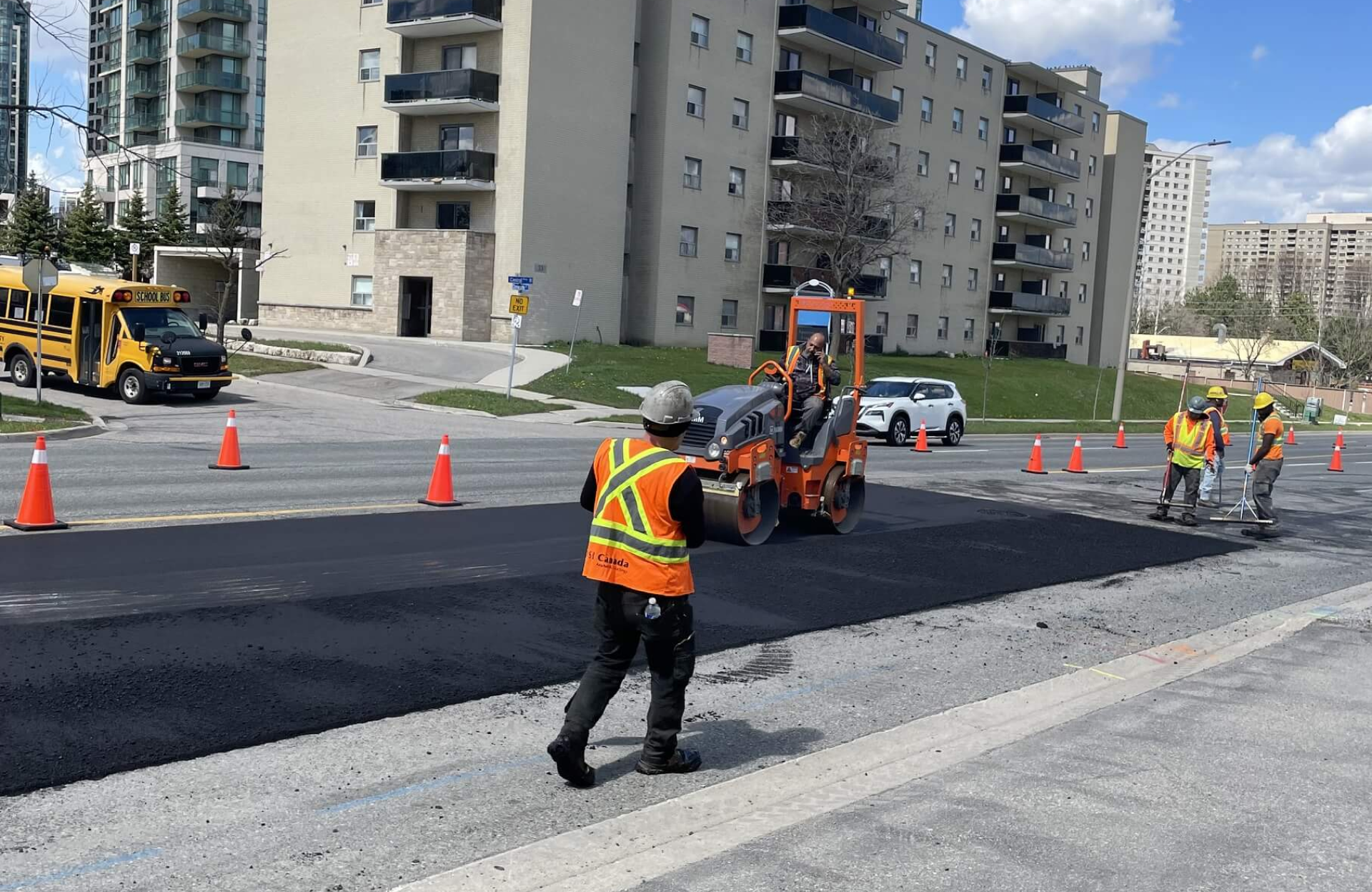 Seal Canada Road construction workers are paving and smoothing asphalt on a street, with orange cones set up for safety near apartment buildings and a school bus, carefully coordinating the sealcoating scheduling to minimize disruptions in the neighborhood. Asphalt & Coatings