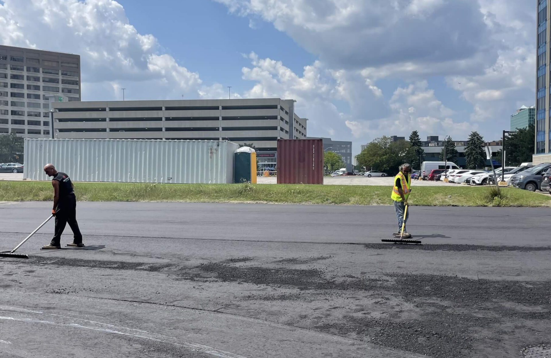 Seal Canada Two workers spread asphalt on a parking lot with brooms under a partly cloudy sky, with office buildings and parked cars in the background. Asphalt & Coatings