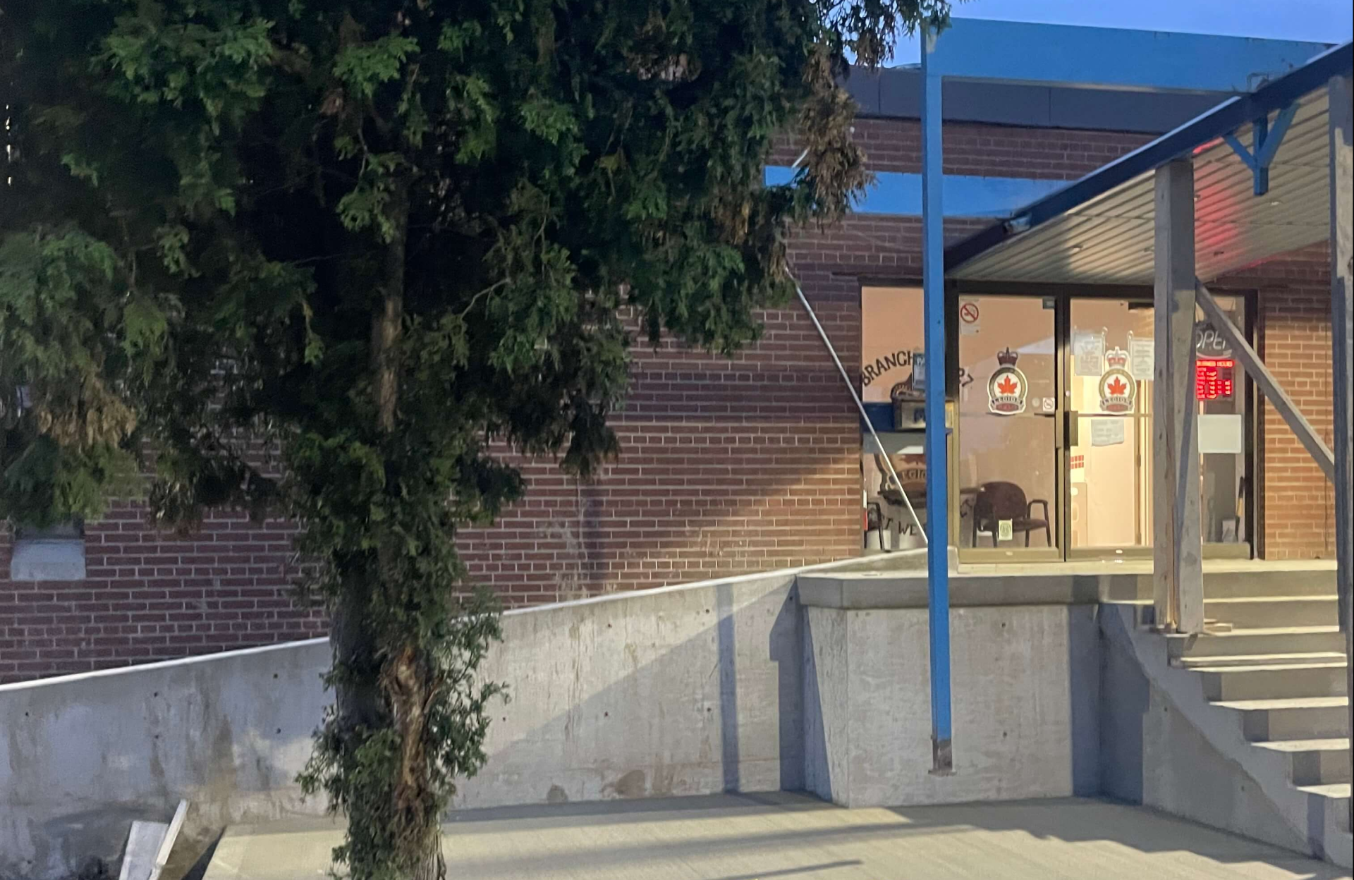 Seal Canada A brick building entrance with glass doors, a covered ramp, and a tree in the foreground—where signs of root damage on the commercial pavement hint at nature’s persistent presence. The area is lit by natural light, suggesting early morning or evening. Asphalt & Coatings