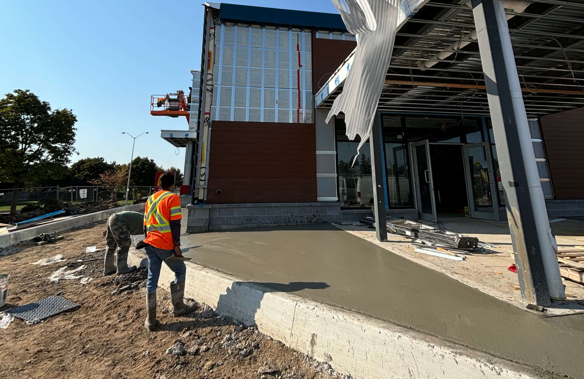 Seal Canada Two construction workers in safety vests smooth freshly poured concrete outside a commercial building under construction on a sunny day, ensuring the site meets high standards for commercial interlock maintenance. Asphalt & Coatings