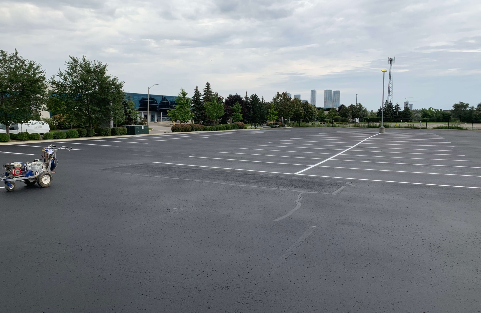 Seal Canada A mostly empty parking lot with freshly painted white lines, some trees and buildings in the background, and a line striping machine on the left—perfectly illustrating the process of expanding an existing commercial parking lot. Asphalt & Coatings