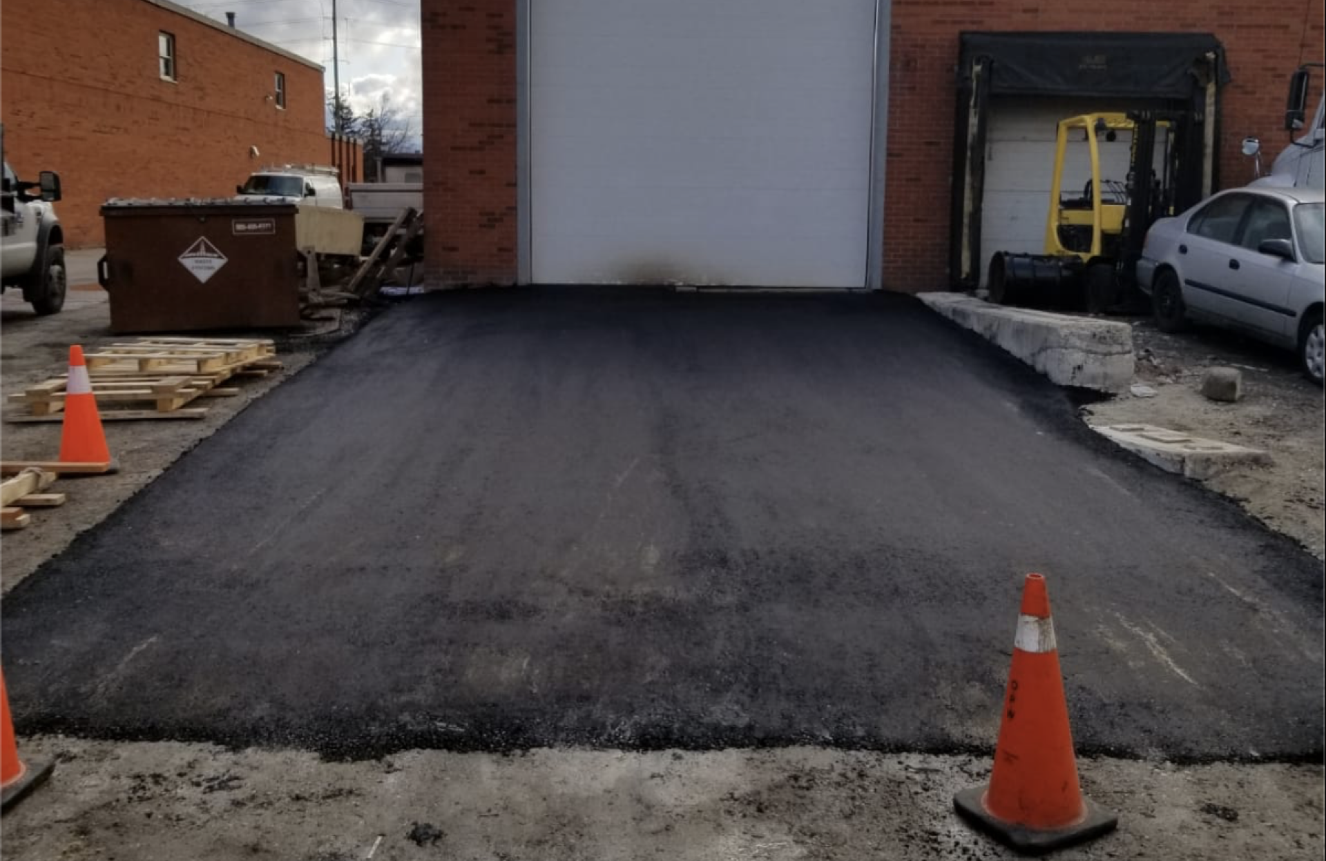 Seal Canada Freshly paved asphalt ramp leading to a warehouse garage door, with orange traffic cones at the entrance and a forklift, dumpster, and car nearby—the surface is protected with durable oil-based sealcoating for added longevity. Asphalt & Coatings