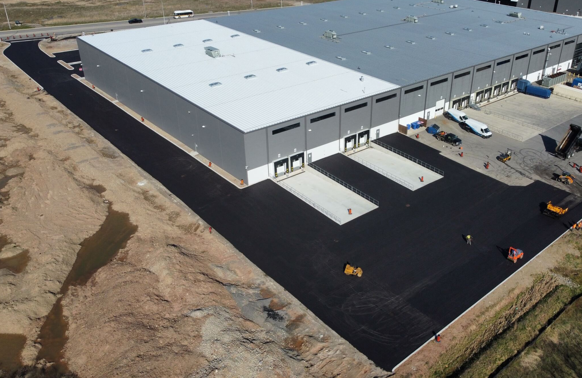 Seal Canada Aerial view of a large industrial warehouse with newly paved black asphalt areas, several vehicles, and construction equipment visible outside—an impressive example of commercial paving projects in action. Asphalt & Coatings