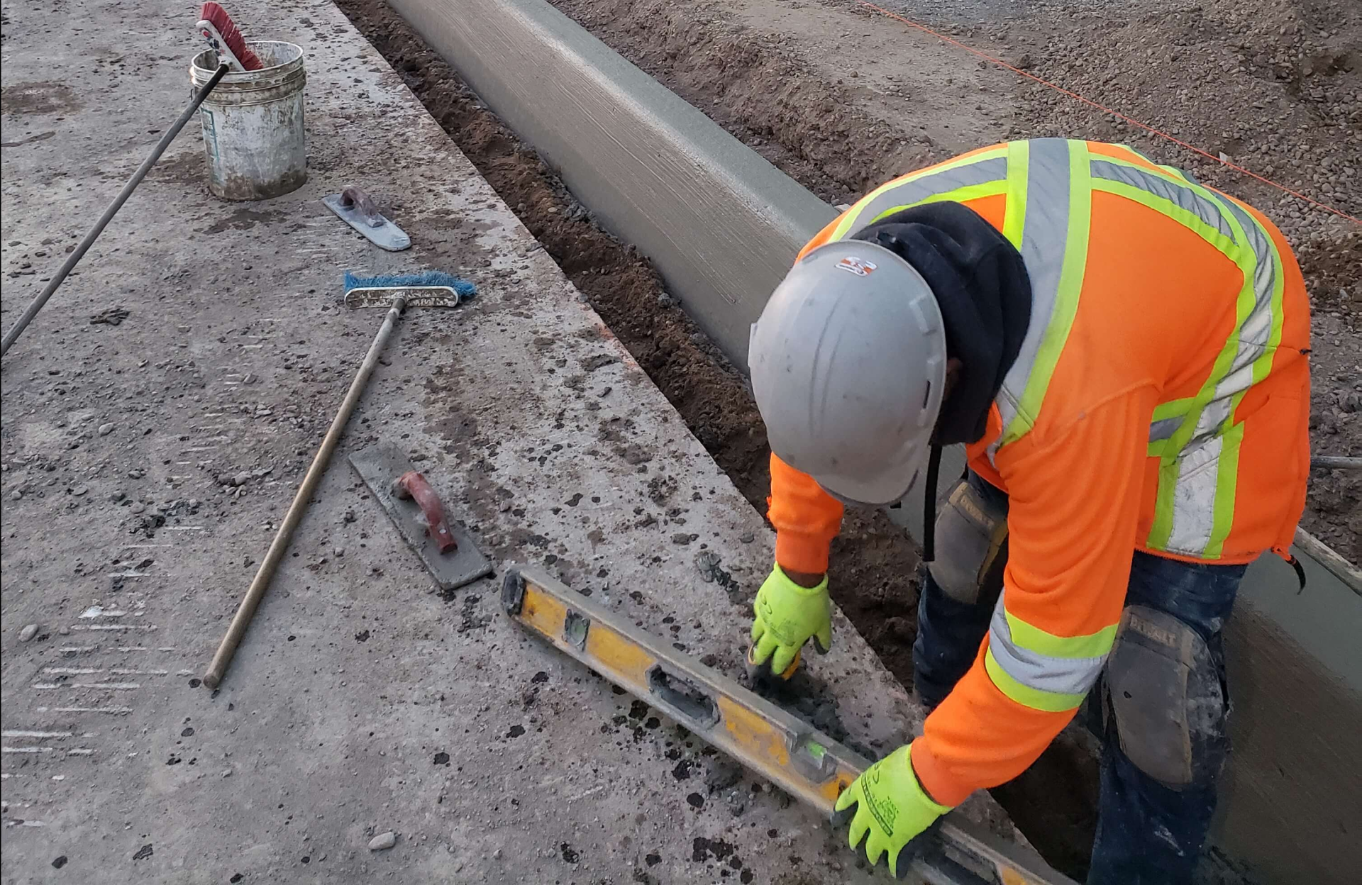 Seal Canada Construction worker in safety gear uses a level tool to check the alignment of a newly built concrete curb, which will soon support an asphalt overlay. Nearby are a rake, a trowel, and a bucket on the ground. Asphalt & Coatings