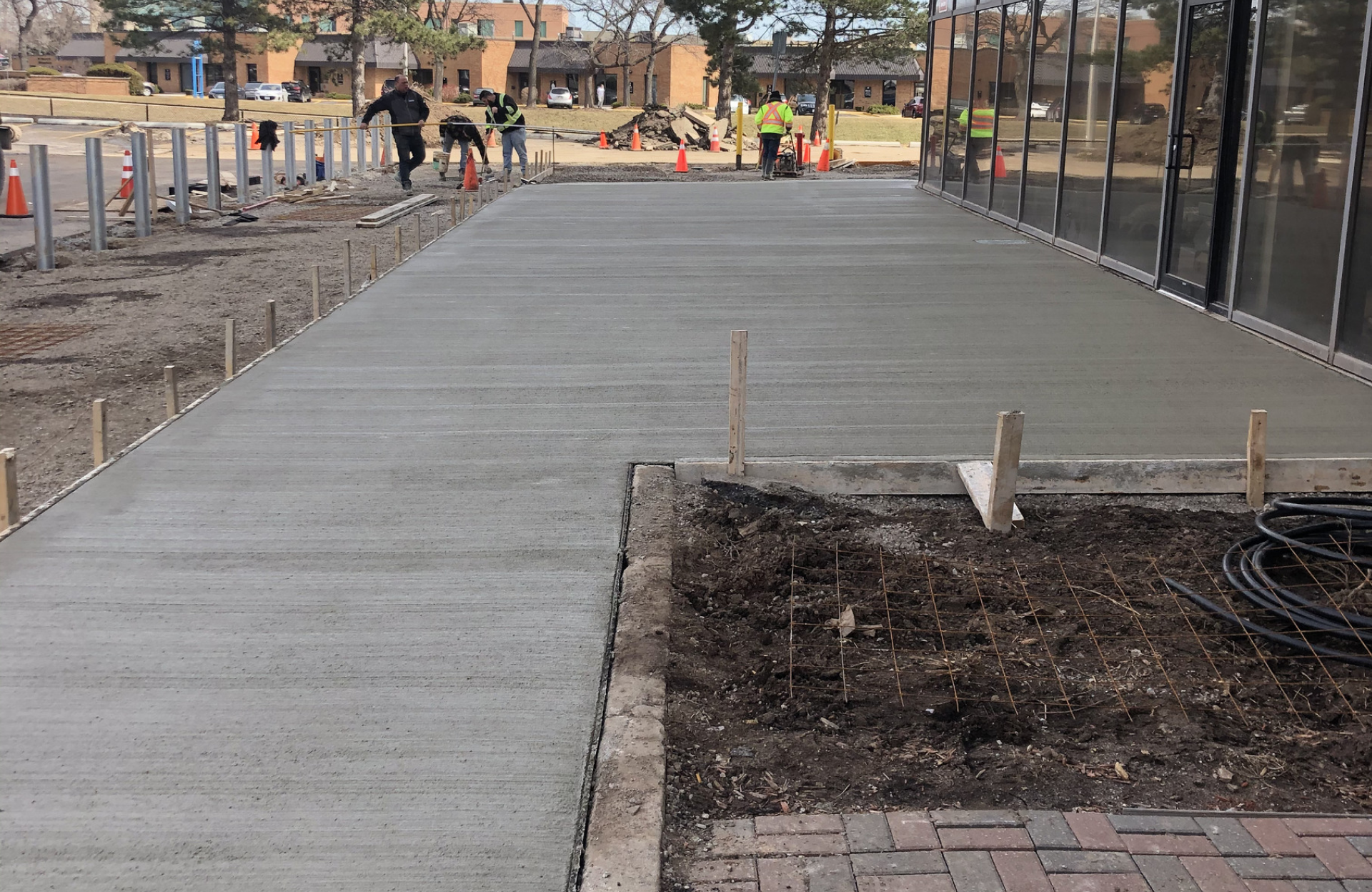 Seal Canada Construction workers smooth a newly poured concrete walkway outside a glass-fronted building, shielded from UV rays as they work away from the nearby asphalt. Asphalt & Coatings