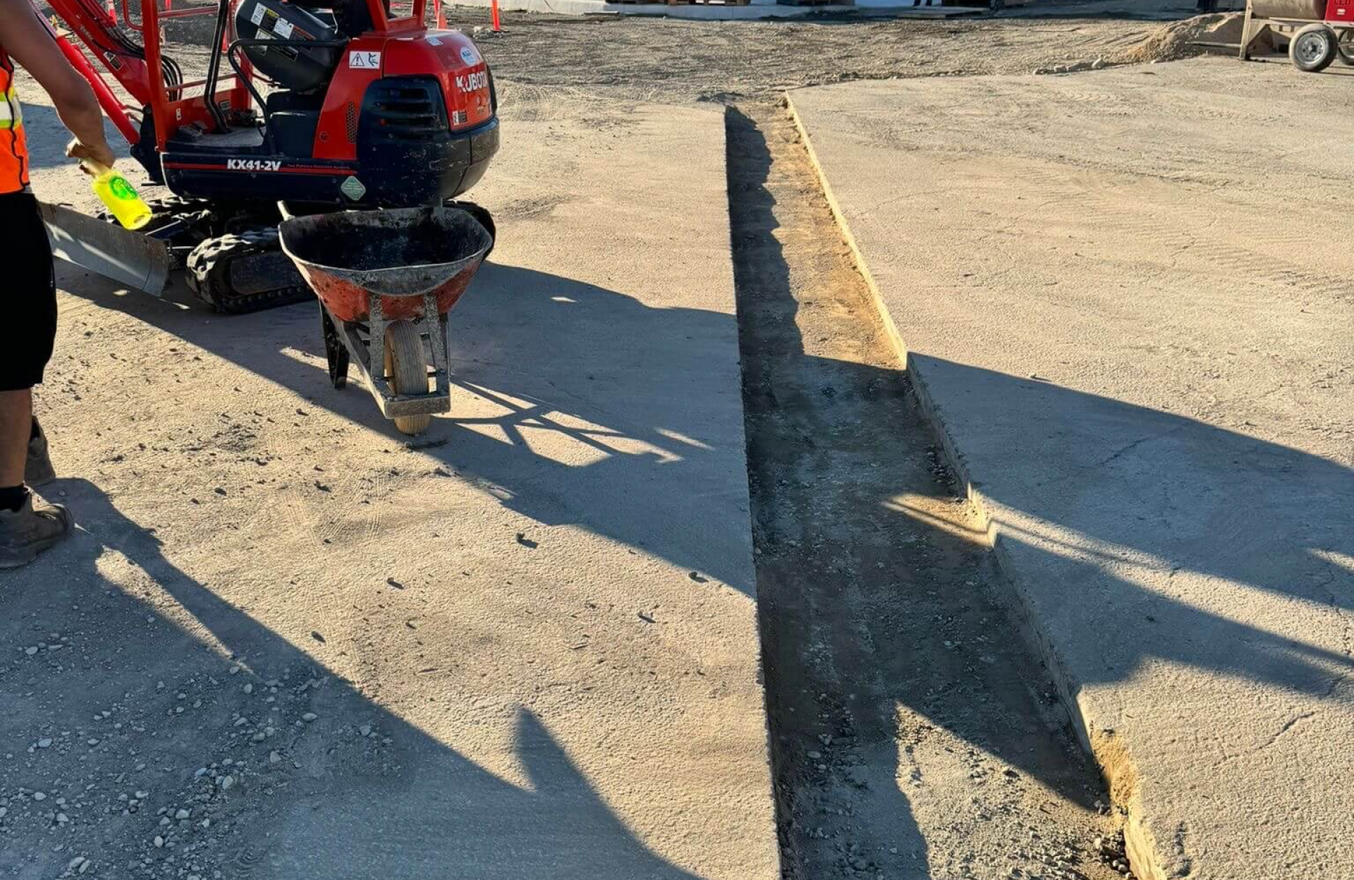 Seal Canada A worker with a wheelbarrow stands next to a small excavator beside a long parking lot drainage trench cut into a concrete surface. Asphalt & Coatings