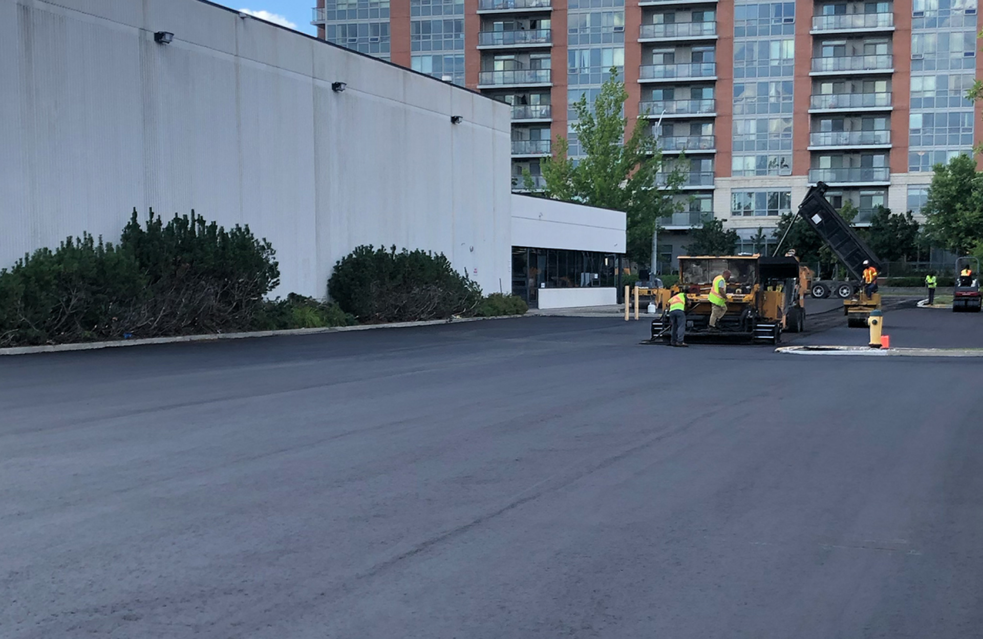Seal Canada Workers operate an asphalt paver on a newly paved parking lot, a key stage in the parking lot lifecycle, in front of a building and apartment complex. Asphalt & Coatings